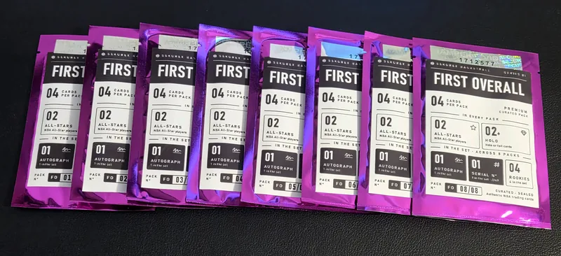 All eight FIRST OVERALL packs fanned out, rose-chrome pouches numbered FO 01/08 to 08/08, each with a holographic tamper-evident tag